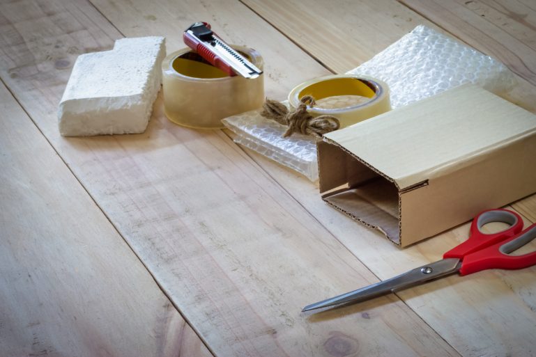 Un petit colis au milieu de divers objets : papier-bulle, paire de ciseaux, rouleau adhésif