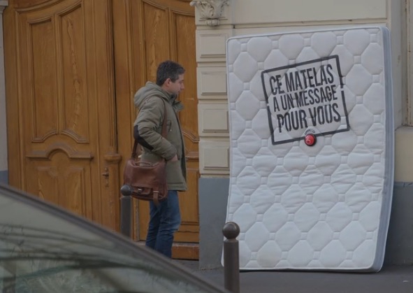 Un homme regarde un matelas posé dans la rue avec la mention "ce matelas a un message pour vous" inscrite dessus