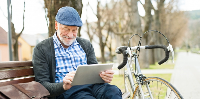 Un senior a posé son vélo et consulte sa tablette sur un banc dans un parc.