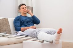 Un homme téléphone assis sur un canapé. Il a la jambe dans le plâtre.