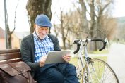 Un senior a posé son vélo et consulte sa tablette sur un banc dans un parc.