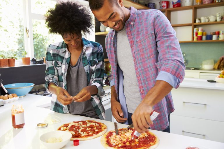 Un couple dans une cuisine. L'homme met de la sauce tomate sur une pizza.
