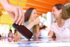 Deux femmes discutent en fond. Au premier plan, une main d’homme subtilise un smartphone.