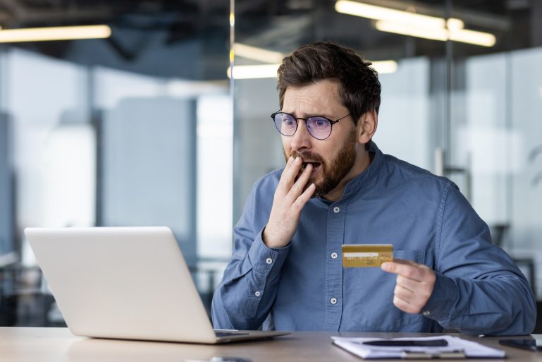 Un homme tenant une carte de crédit à la main a l’air choqué par l’écran